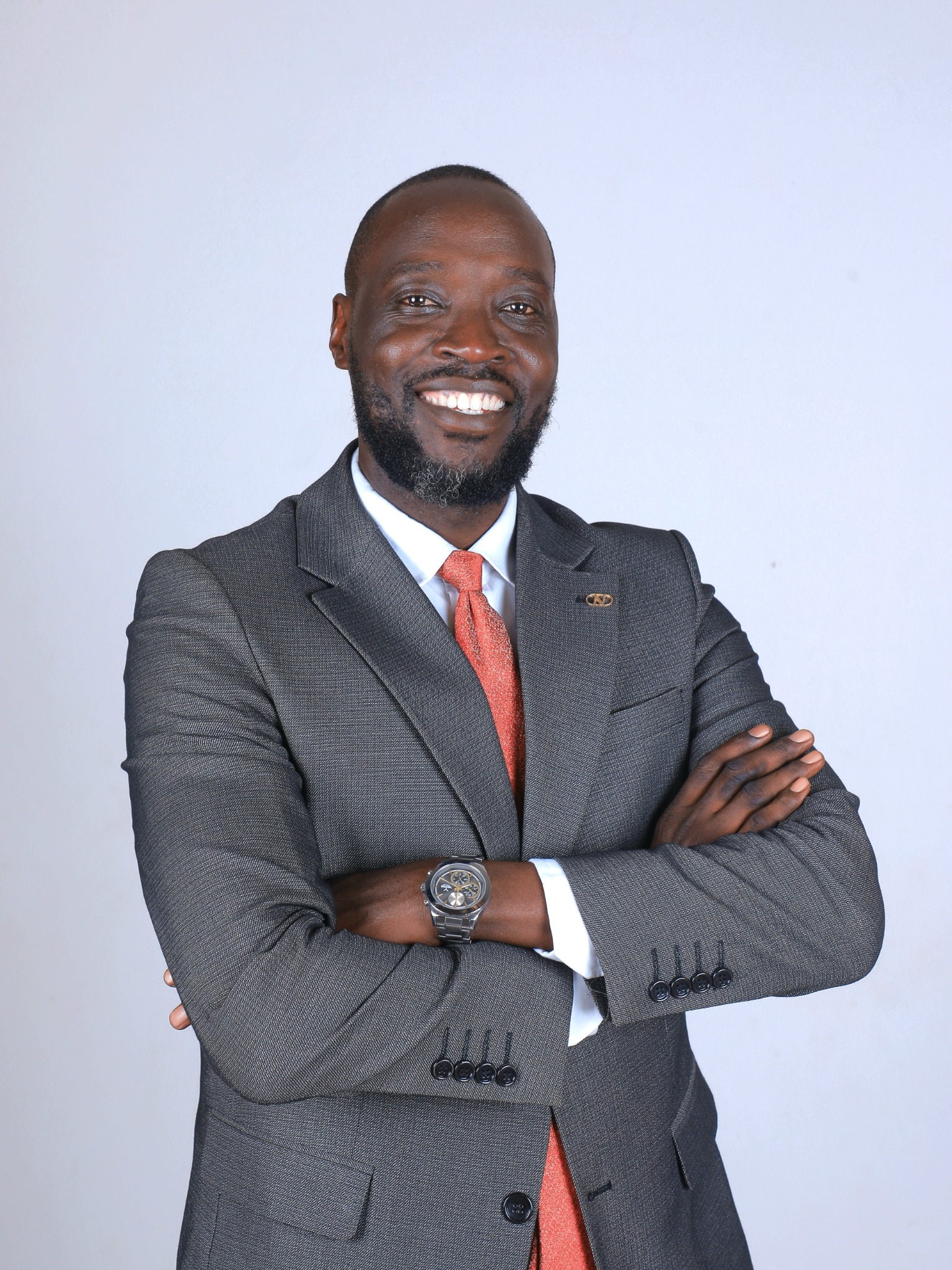 Jerald Onyango Okwera Okuka with arms crossed, wearing a grey suit and red tie